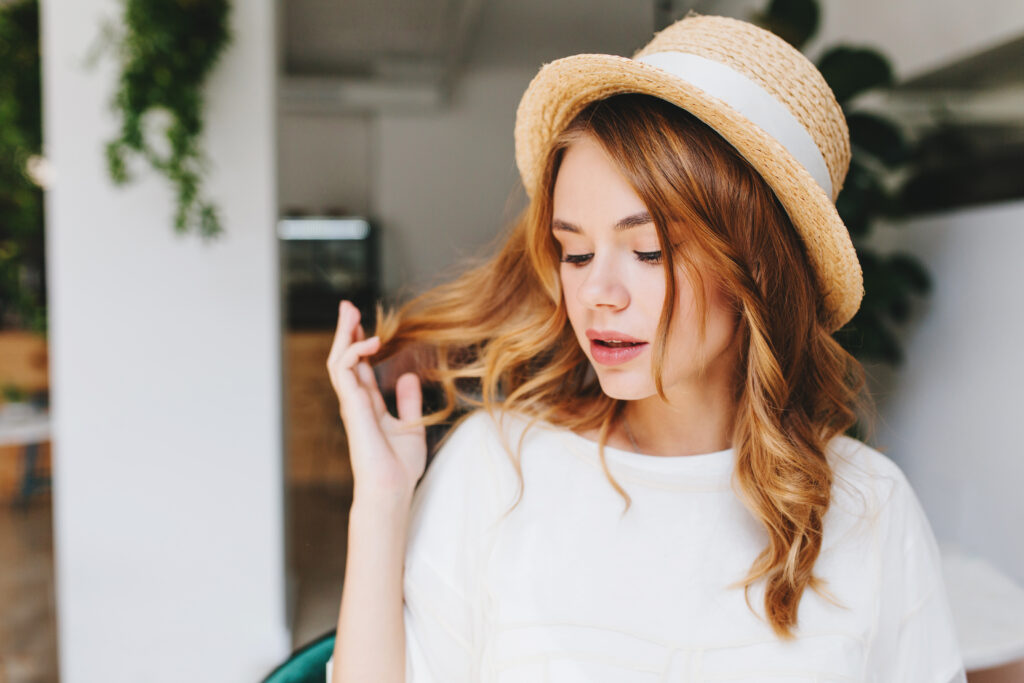 Close-up portrait of dreamy young lady with curly hairstyle and pale skin wearing elegant straw hat decorated with white ribbon. Indoor photo of cute girl with eyes closed plays with blonde hair..