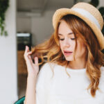 Close-up portrait of dreamy young lady with curly hairstyle and pale skin wearing elegant straw hat decorated with white ribbon. Indoor photo of cute girl with eyes closed plays with blonde hair..