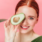 Close up view of gorgeous ginger woman with avocado. Studio shot of excited caucasian girl with healthy food isolated on pink background