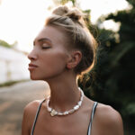 Pretty tanned blonde woman with hair bun and pearl necklace poses with closed eyes outside. Charming girl in top walks outdoors