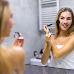 Beauty, make up, cosmetics, morning and people concept – young woman with makeup brush and contouring and highlighting palette looking to mirror at home bathroom